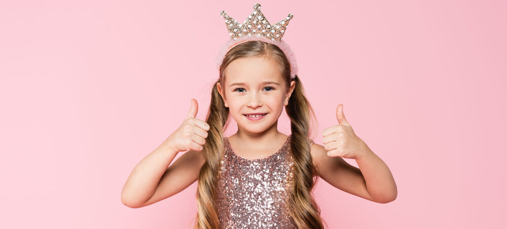 cheerful little girl in dress and crown showing thumbs up e1618726054415